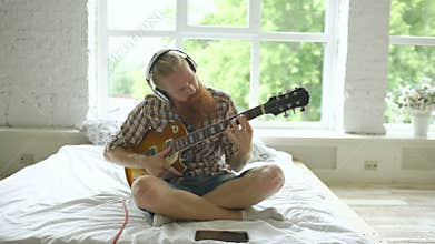 Attractive bearded man in headphones sitting on bed learning to play guitar using tablet computer in modern bedroom