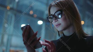 Charming blonde in a good mood in the glasses with black rim and dark clothes is on the evening street. She is near the
