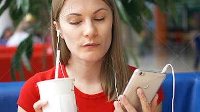 Woman in red t-shirt sitting in cafe using her smartphone, listening music drinking cola from paper cup