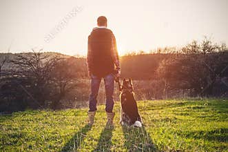 A man with a beard walking his dog in the nature, standing with a backlight at the rising sun, casting a warm glow and