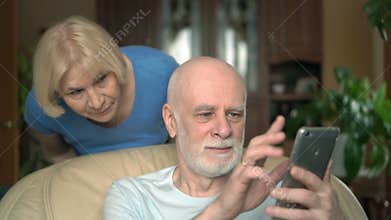 Senior couple at home. Husband using smartphone. Wife spying on her husband from his shoulder