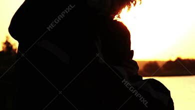 Mother hugging her son near the river at sunset silhouetted dawn, glare on water