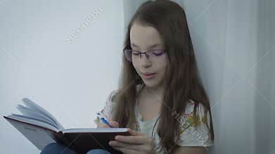Emotional little girl with glasses writes in his diary at window.