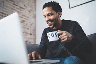 Cheerful African man using computer and smiling while sitting on the sofa.Black guy holding ceramic cup in hand.Concept