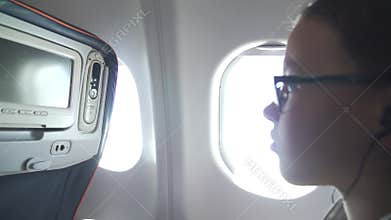 Young girl with glasses and headphones watches video on the monitor built into armchair in the cabin of the airplane