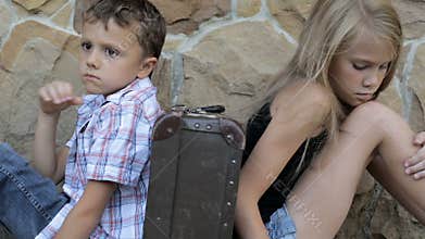 Sad brother and sister are sitting by the wall with a suitcase at the day time