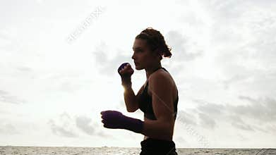 Tough pretty woman shadow boxing. Beautiful female boxer training by the sea on the beach in the morning, throwing