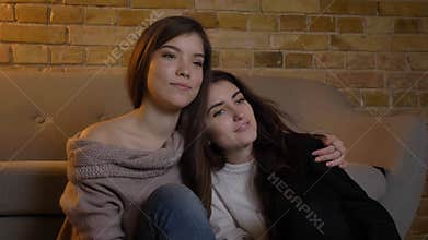 Closeup portrait of two young pretty women hugging and watching TV in a cozy apartment indoors