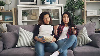 Bored young women are watching TV together at home and eating popcorn sitting on sofa in living room. Asian girl is