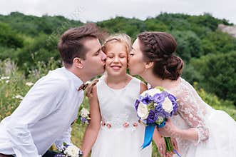 Young parents in wedding dresses kiss their young daughter in cheeks