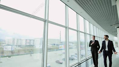Two businessmen chat together as they walk along through a busy modern office building