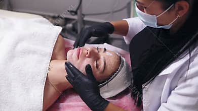 Young woman with blemish skin lying on the couch while professional cosmetologist is applying face cream and making face