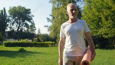 Delighted aged man walking in the park with an exercise mat in his hands