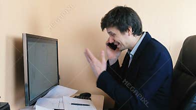Frustrated man working at the computer and calling mobile phone. Compares graphics on the screen and on paper.
