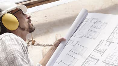 Architect man at work, wearing helmet and headphones looking at blueprint, check the construction house project plan, in