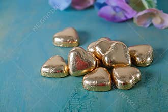 Heart shaped chocolates in golden foil on an old green table. Blurred background with flowers. Wedding card. Soft focus.