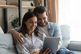 Smiling spouses hug on sofa watch wedding video on pad