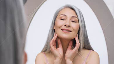 Gorgeous Asian mature woman looking at mirror touching skin.