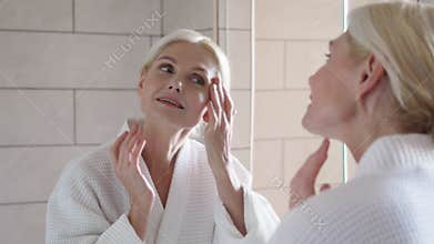 Happy smiling mid age blond woman standing looking at mirror. Skincare concept.
