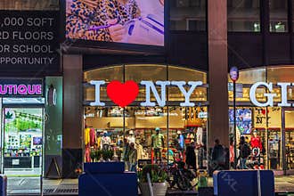 Storefront with large \"I Love NY\" sign illuminated at night in New York City, with people walking by and shopping.