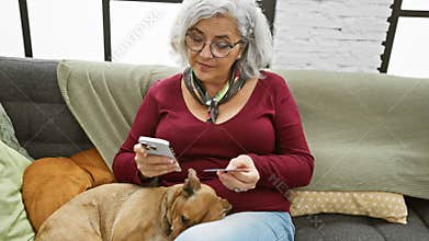 Mature woman using smartphone and credit card for online payment, with her dog beside her on the indoor couch