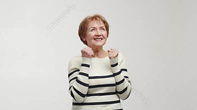 An elderly positive, confident and pleased woman stands, says yes, looks up, smiles, clenches hands into fists and joins