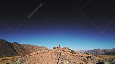 A white man wearing FPV goggles and a helmet sits in the mountains and controls an FPV acrodrone quadcopter flying