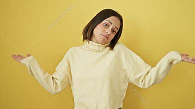Clueless young hispanic woman stands doubted, her beautiful face alone against a yellow wall, hands and arms raised in confusion,