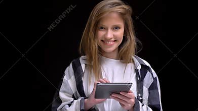Portrait of young caucasian long-haired girl in shirt working with tablet and smiling into camera on black background.