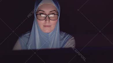 Portrait of Muslim woman`s face in hijab and glasses at night in the dark uses computer