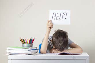 Sad tired frustrated boy sitting at the table with many books and holding paper with word Help. Learning difficulties