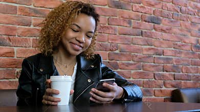 A young modern beautiful African-American girl is smiling talking on the phone and drinking a drink from a white cup