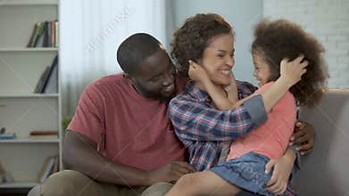 Mother tickling and amusing her little daughter, family sitting on couch