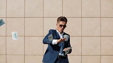 Young happy businessman scatter the dollars and funny dancing in the street. He walking near the office center, slow