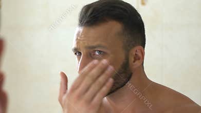 Unhappy man checking wrinkles on face in front of mirror in bathroom, aging