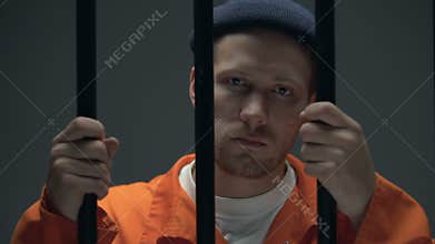 Imprisoned male holding bars and looking to camera, feeling guilty and desperate