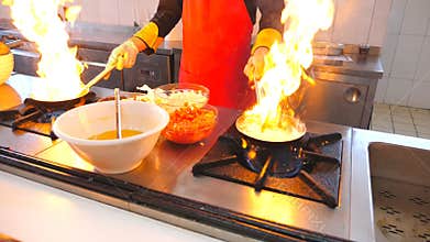 Professional chef holding two pans and cooking flambe style dish in modern kitchen of restaurant. Male cook frying