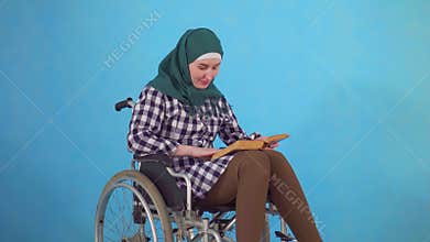 Young Muslim woman disabled in wheelchair with book in hand on blue background