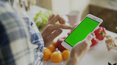 Closeup of woman`s hand browsing smartphone with green screen on kitchen at home