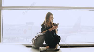 Happy woman sits with smartphone by airport window. Caucasian girl with backpack using messenger app in terminal. 4K.