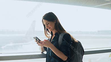 Young girl using smartphone near airport window. Happy European woman with backpack uses mobile app in terminal. 4K.