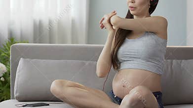 Future mother sitting on couch in headphones, stretching upper body, bodycare