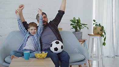 Family members father and son are watching soccer match on TV at home, cheering, celebrating victory and eating snacks