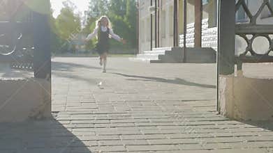 A little funny schoolgirl in school uniform and a briefcase on her shoulders runs out of school