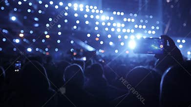 Audience with hands raised at a music festival and lights streaming down from above the stage. A lot of people with