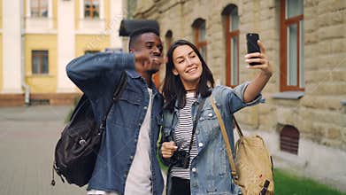 Happy tourists friends with backpacks are taking selfie using smartphone showing hand gestures v-sign and thumbs-up