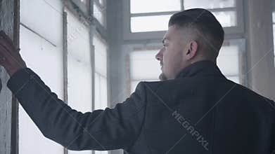 Handsome bearded young man with serious face touches glass of windows.