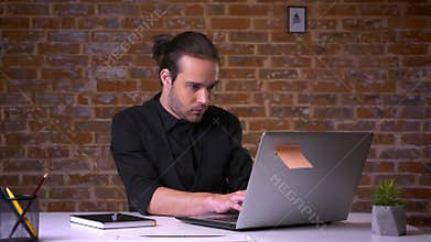 Good looking caucasian office worker thinking and biting finger in front of the computer with brick wall behind