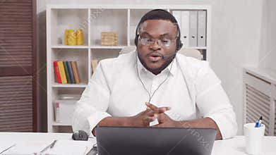 Workplace of freelancer. African-American man works at home office using computer and headset. Employee is having a