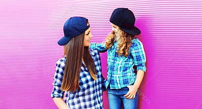 Portrait of beautiful young mother with little girl child wearing checkered shirts, baseball cap in city on pink background